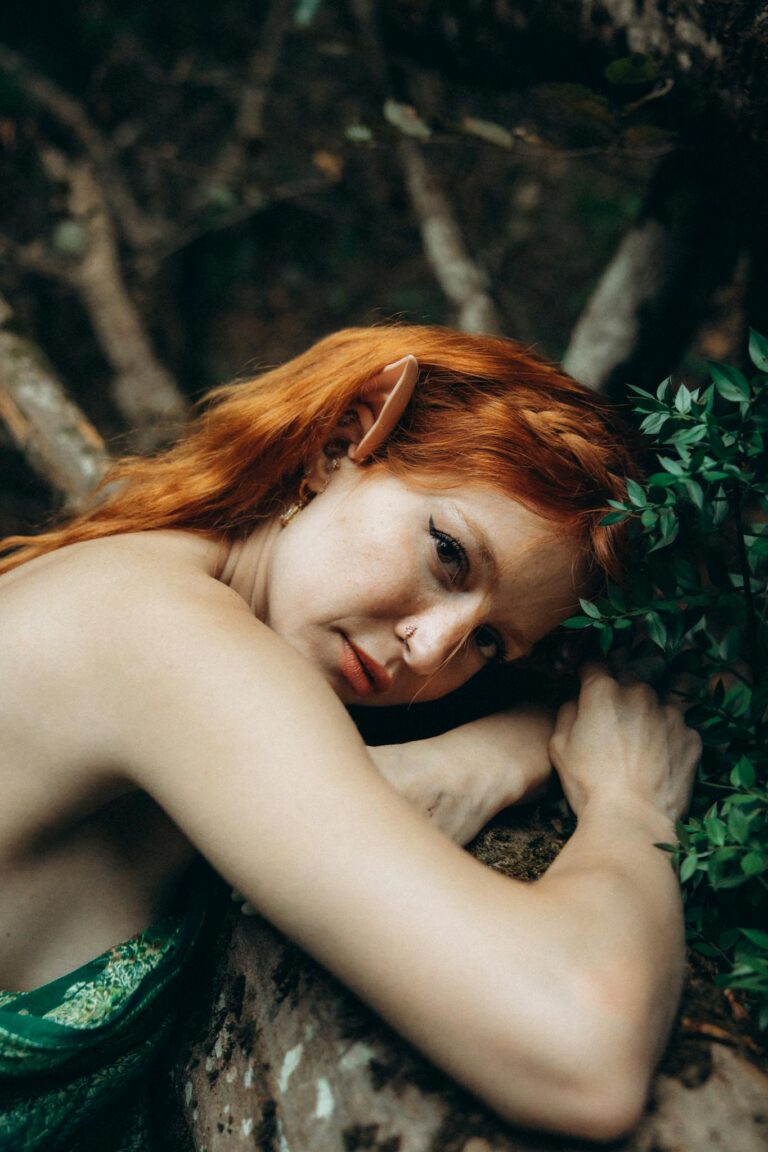 A woman with red hair and green dress leaning against a tree